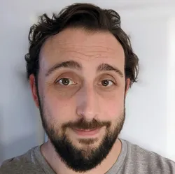 A man with short, dark, wavy hair and a beard is looking at the camera. He is wearing a gray shirt and standing against a plain background.